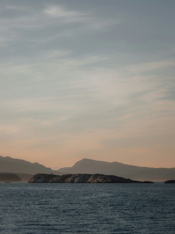 Calm sea with a small rocky island and distant mountains at sunset.