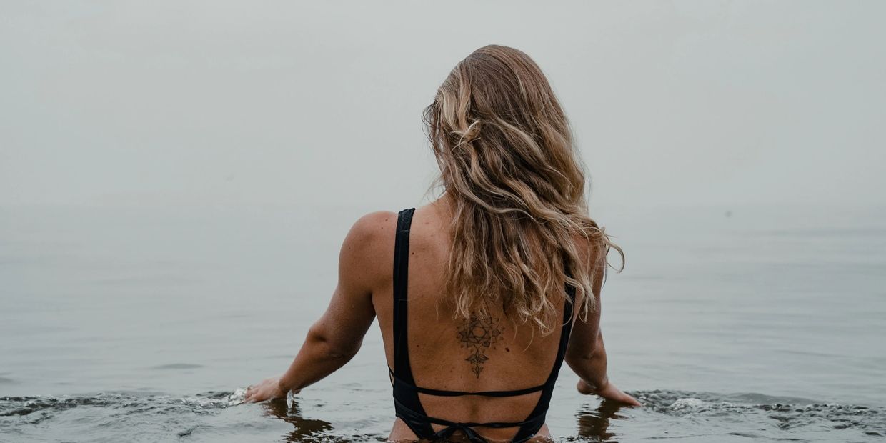 Woman with tattooed back in black swimsuit wading into calm water.
