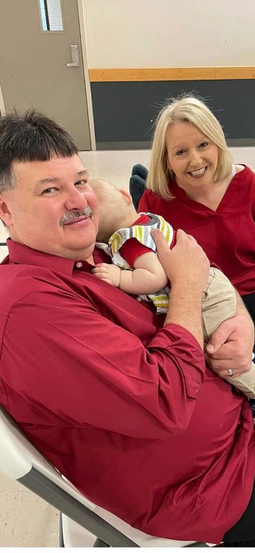 Man in red shirt holds a baby while a woman smiles nearby.