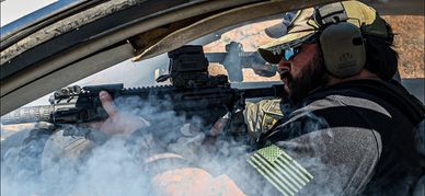 Man shooting a rifle from a car window with smoke around.