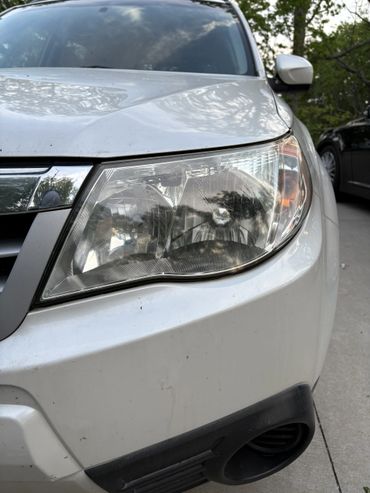 Close-up of a white car's dirty headlight and front bumper.