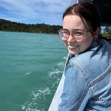 A smiling young woman with glasses on a boat near green water and forested shore.