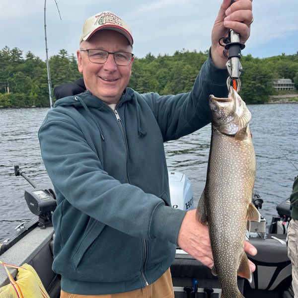 Man holding a large fish on a boat with a lake and trees in the background.