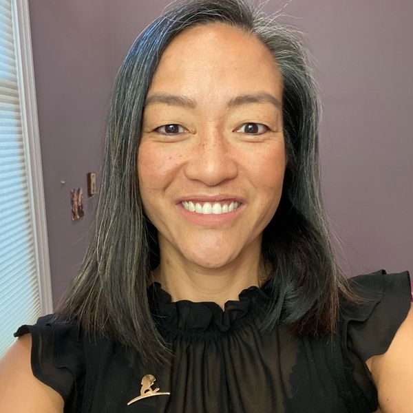 A smiling woman with gray-streaked black hair wearing a black top and a pin.
