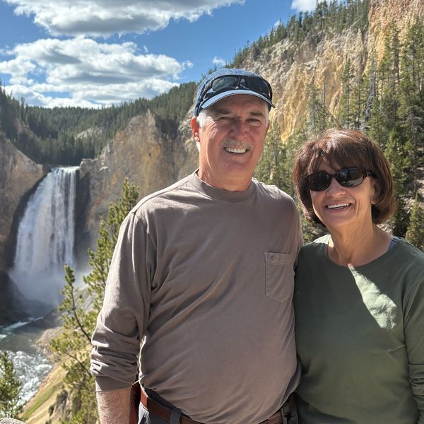 Smiling couple posing near a scenic waterfall with forested cliffs in the background.