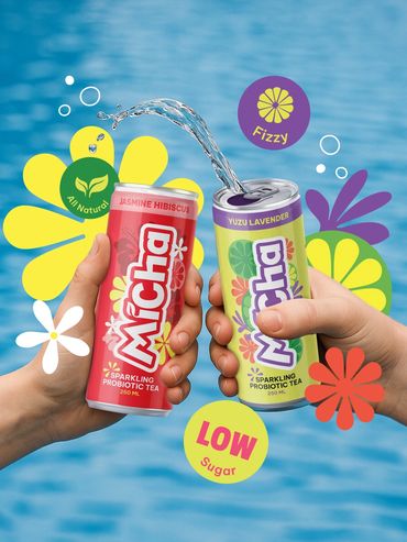 Two hands holding colorful cans of Micha sparkling probiotic tea by water.