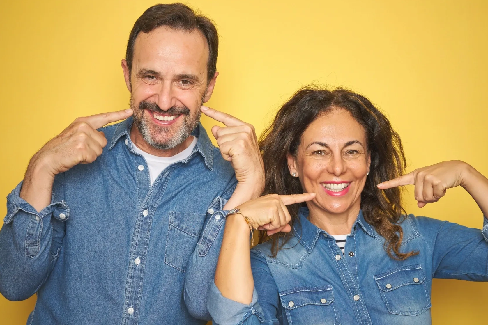 A smiling man and woman in denim shirts pointing at their teeth against a yellow background.