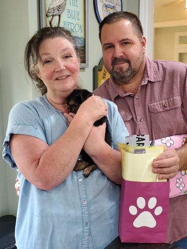 Smiling couple holding a small puppy and a gift bag with a paw print.