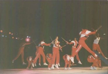 Group of performers in red outfits mid-air with sticks on stage.