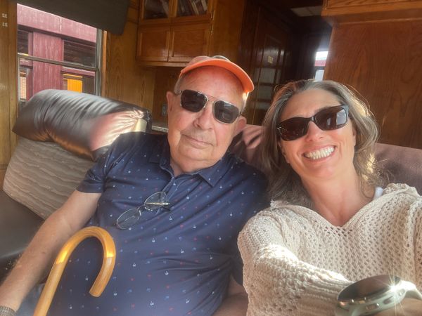An elderly man and a woman wearing sunglasses, sitting together indoors, smiling.