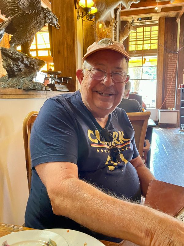 Smiling elderly man wearing glasses and a cap sitting in a rustic restaurant.