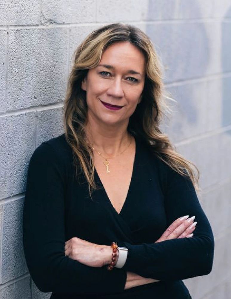 Confident woman with crossed arms leaning against a gray brick wall.