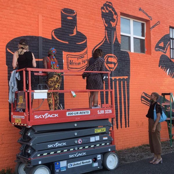 Three people on a lift painting a vibrant mural on an orange brick wall while another person watches.