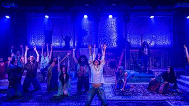 A theatrical performance with a vibrant ensemble cast on stage under blue lighting.