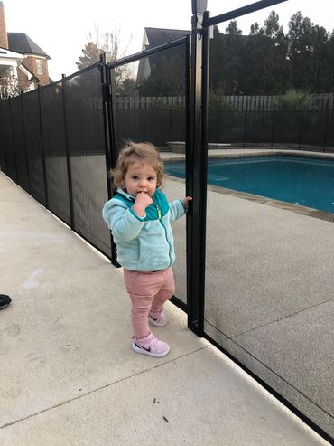 Toddler standing by a pool safety fence with a finger in their mouth.