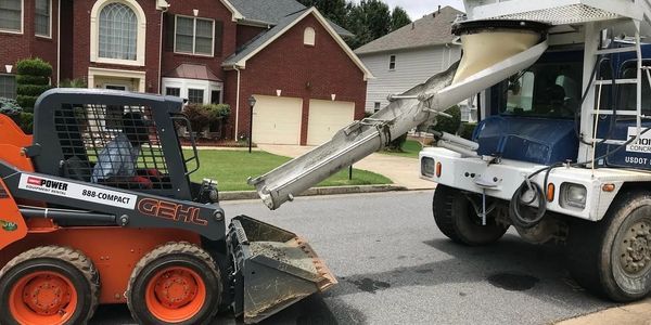 A compact skid steer loader receiving concrete from a mixer truck on a suburban street.