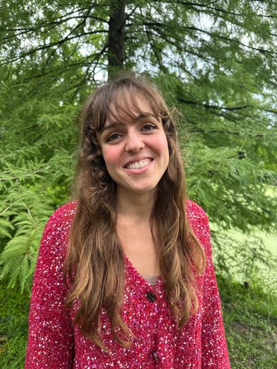 A smiling woman in a red sweater stands outdoors with green trees in the background.