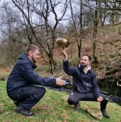 TWO MEN IN NATURE CROUCHING, ONE HOLDING A TIBETAN SINGING BOWL