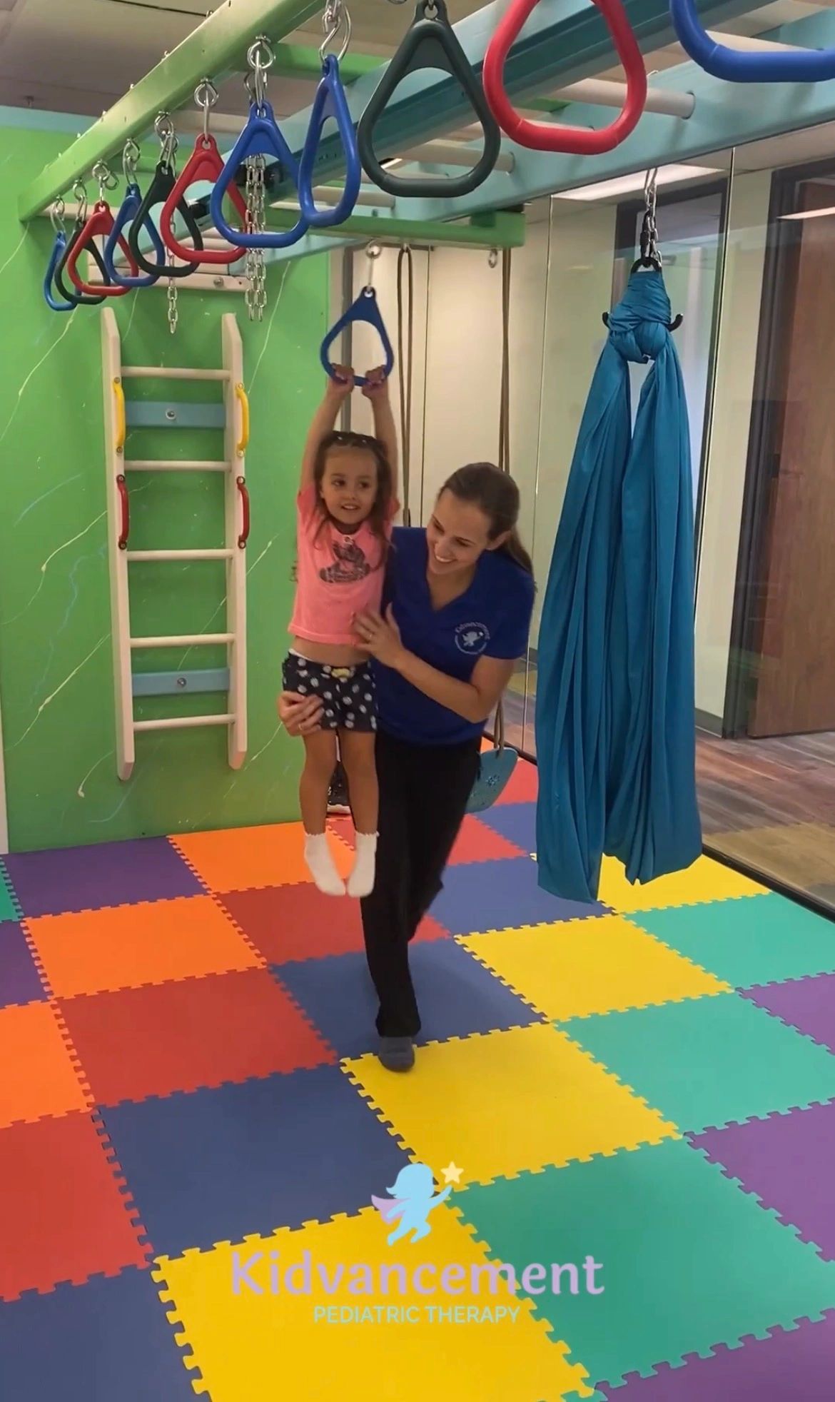 Therapist helping a child hang on rings in a colorful pediatric therapy room.