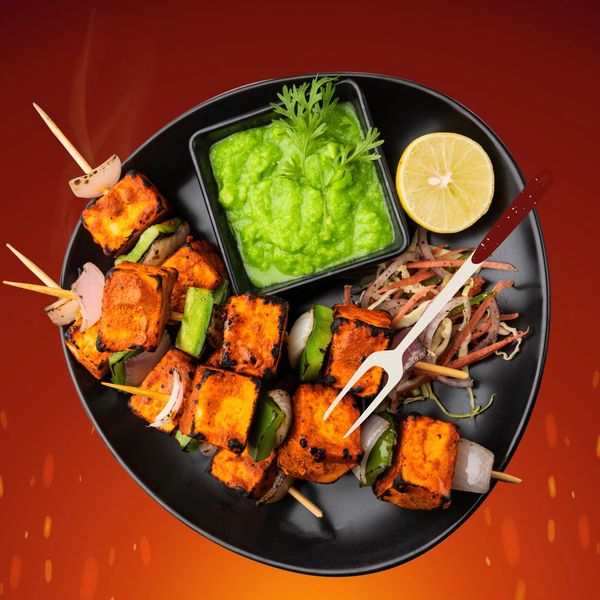 Grilled paneer tikka skewers with green chutney and lemon on a black plate.