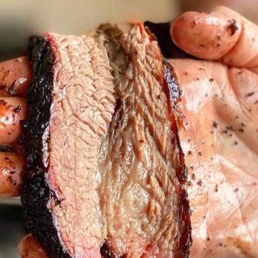 Close-up of a hand holding two juicy slices of smoked brisket with a dark bark.
