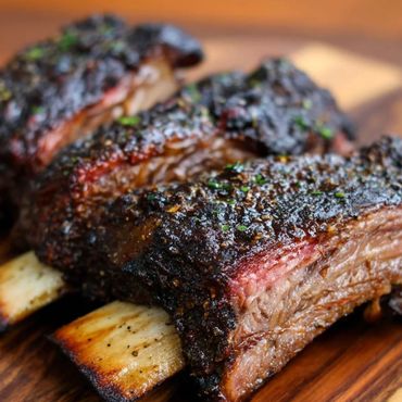 Close-up of smoked beef ribs with a dark crust on a wooden board.