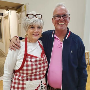 An elderly woman in a red checkered apron and a man in glasses smile together indoors.