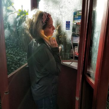 woman person on the phone in a phonebooth, outside with plants and in the rain, making a call receivr