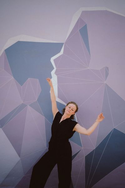 Rayni with eyes closed, arms up in fluid dance pose before purple mural, embodying rest and renewal.