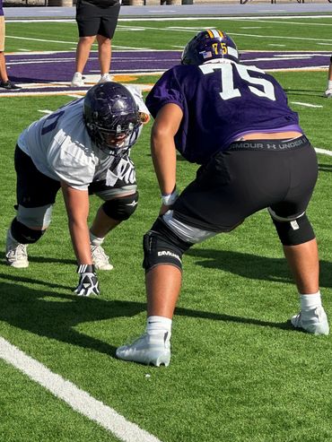 Two football players face off in a practice drill on a sunny turf field.