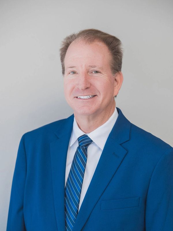 Smiling middle-aged man in a blue suit and striped tie against a plain background.