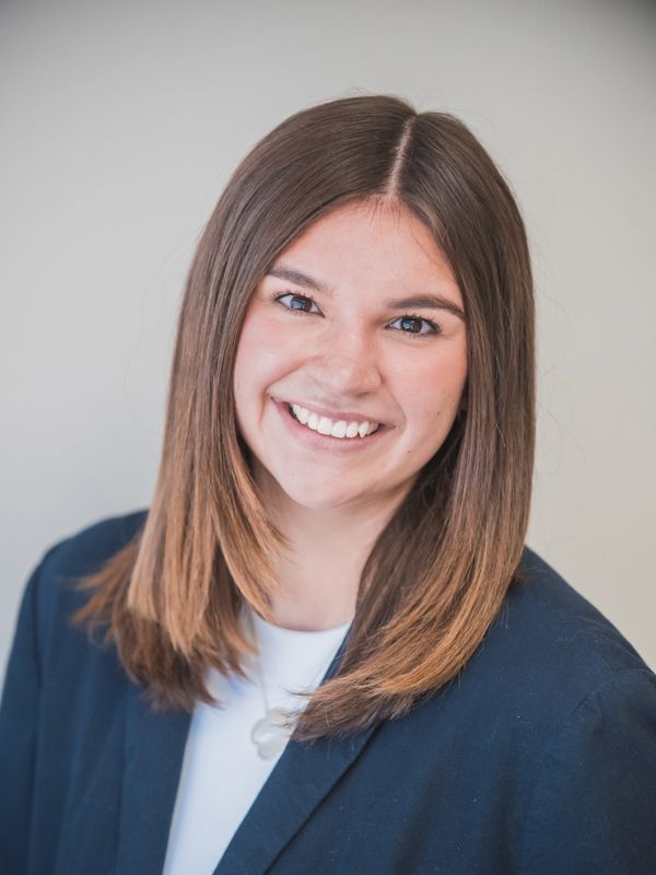 Young woman with straight brown hair smiling warmly, dressed in business attire.