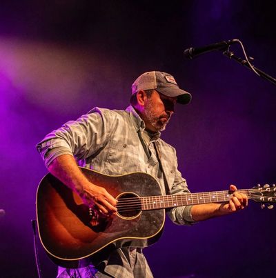 Man playing guitar on stage with purple lighting.