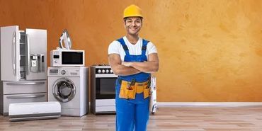 A confident technician in blue overalls and a yellow helmet stands among home appliances.