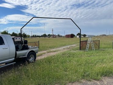 Pickup truck and ladder near a metal gate entrance to a grassy field under a cloudy sky.