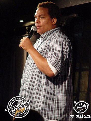 JP JUSTICE performing stand-up on stage in a plaid shirt, holding a mic mid-set at a comedy venue.
