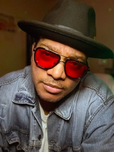 JP JUSTICE in red sunglasses, wide-brim hat, and denim jacket, staring confidently into the camera.