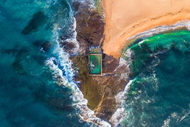 Aerial view of a coastal rock pool surrounded by ocean waves and sandy beach.
