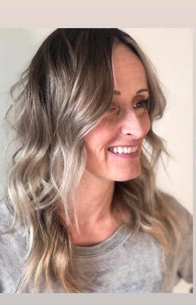 Smiling woman with wavy, highlighted hair and a cozy sweater.