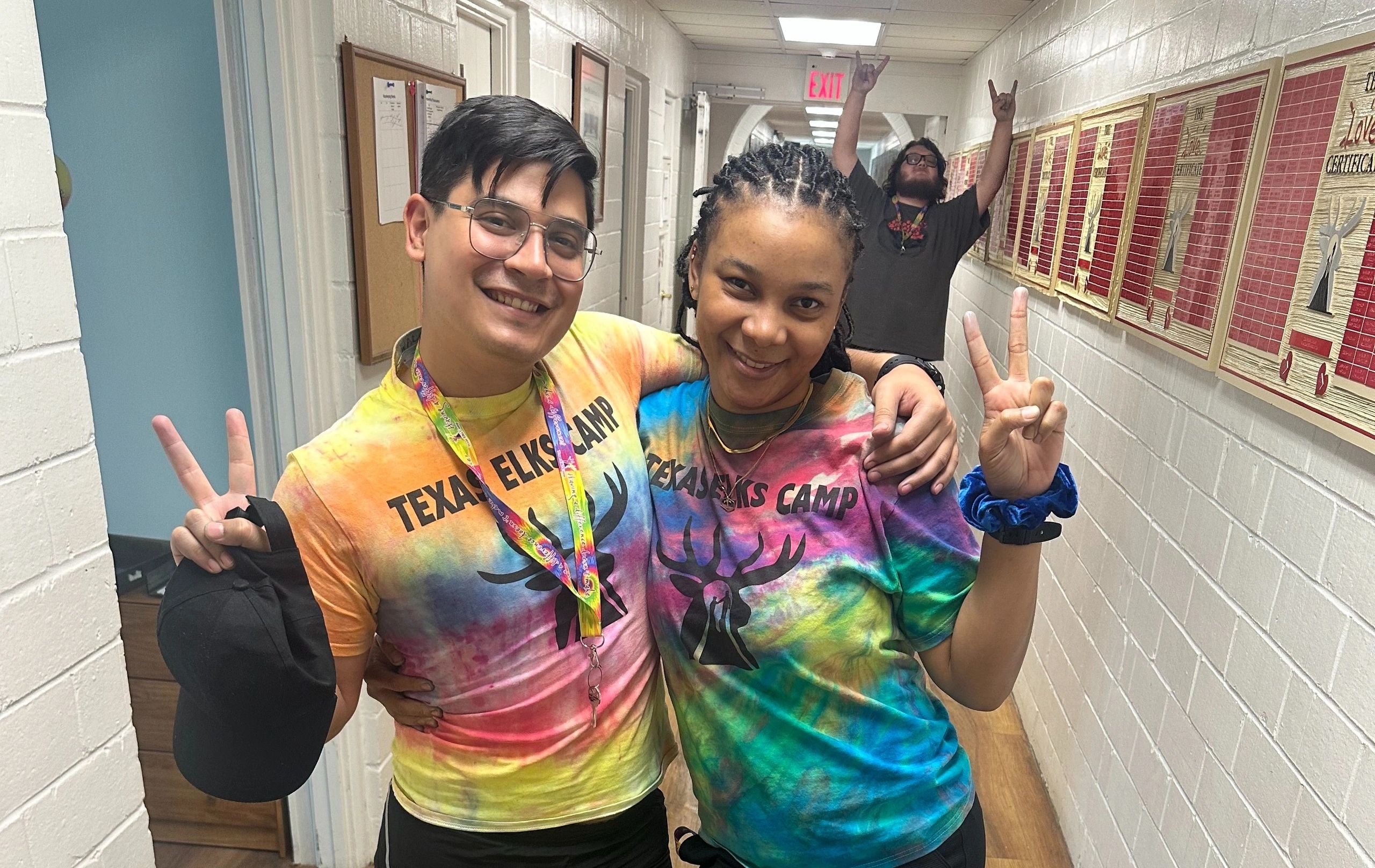 Two people in colorful Texas Elks Camp shirts pose with peace signs, while a third person joyfully raises hands in the background.