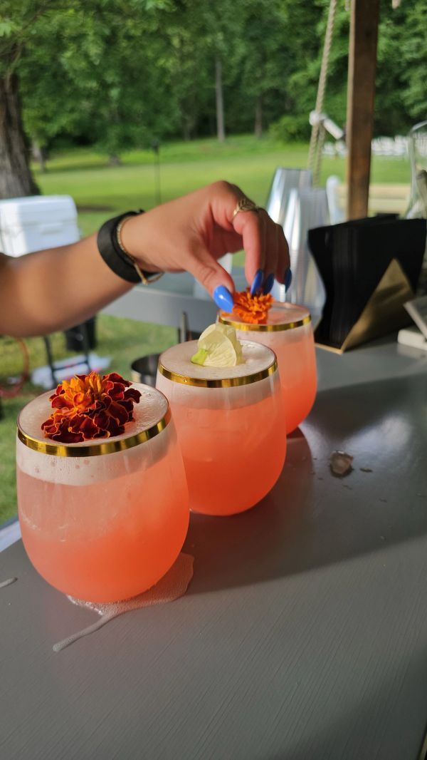 Hand placing flower garnish on pink cocktails in gold-rimmed glasses.