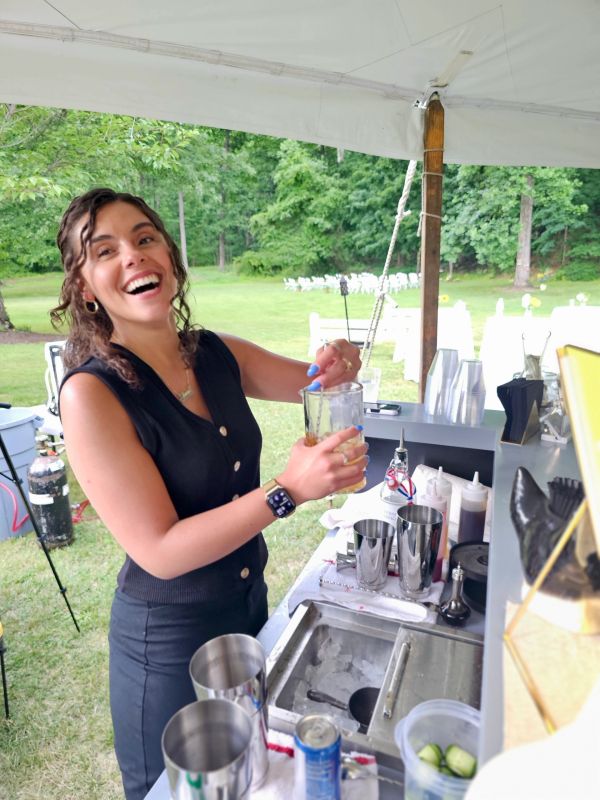 Smiling woman preparing a drink outdoors under a canopy.