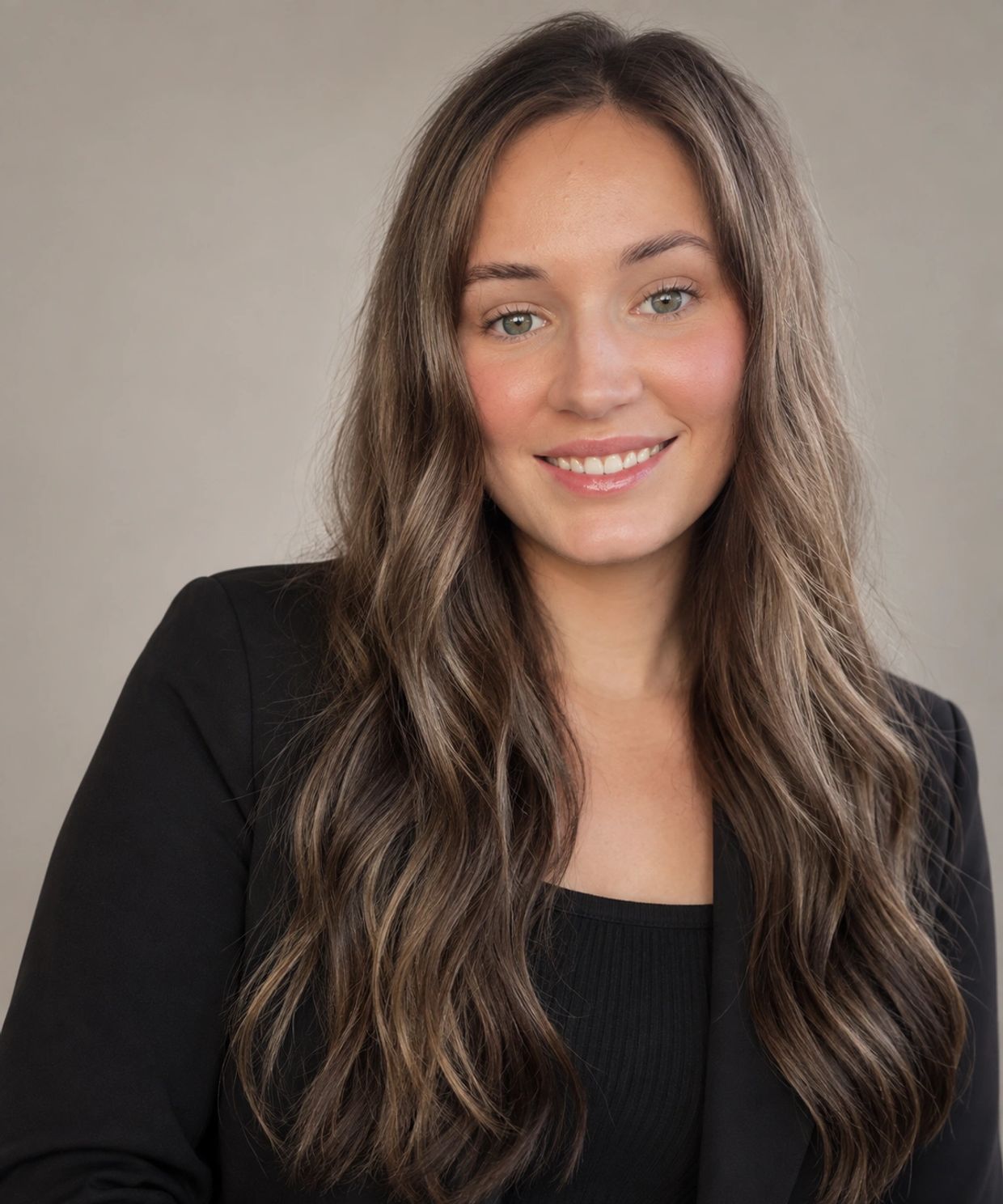 Young woman with long wavy brown hair and a black blazer smiles.