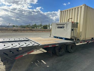 Flatbed trailer carrying a container and an Ingersoll Rand machine under a partly cloudy sky.