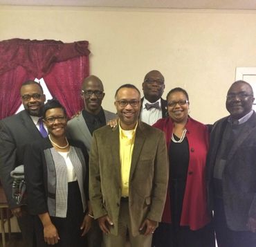 A group of seven adults smiling indoors, dressed in formal and semi-formal attire.