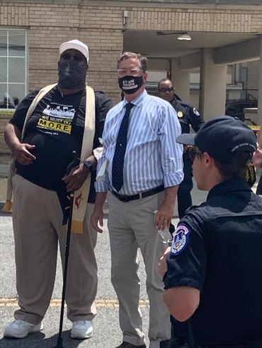 Two men wearing masks stand outdoors, one holding a cane, with police officers nearby.
