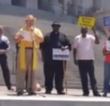 People standing on steps, one speaking into a microphone, others holding signs.