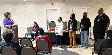 A speaker addresses a group with four people standing and audience seated in a conference room.