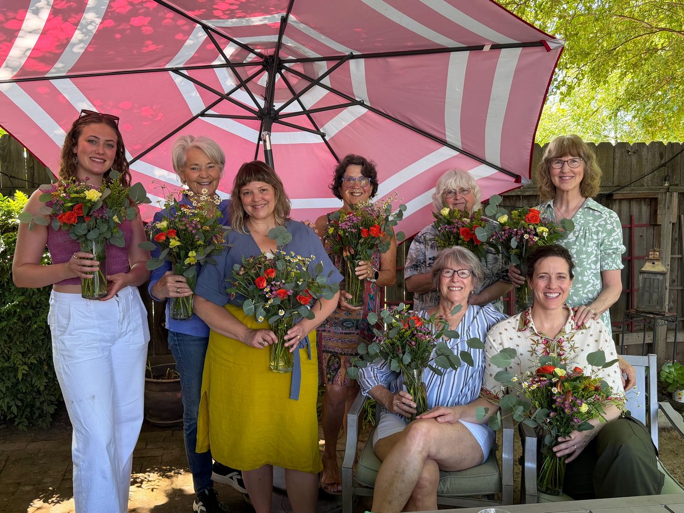 Eight women holding flower bouquets under a large pink umbrella in a garden setting.