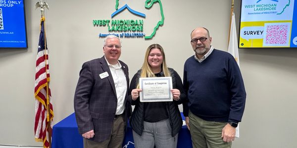 Three people pose for a photo, with the center person holding a certificate at West Michigan Lakeshore Association of Realtors.